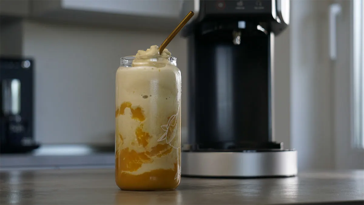 Ein Mango-Matcha-Frappé ist in einem Glas mit Strohhalm angerichtet, im Hintergrund steht die Ninja Creami Deluxe.