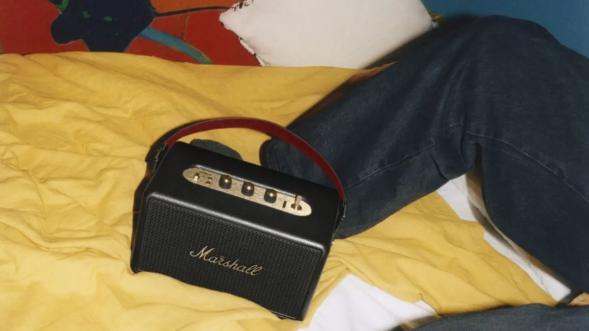 Ein schwarzer Marshall-Lautsprecher mit rotem Griff liegt auf einem gelben Bettlaken neben einer Person in Jeans.