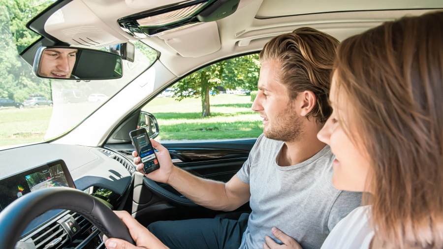 Im Auto hält ein Mann ein Smartphone, während eine Frau daneben sitzt. Ein Rückspiegel zeigt den Fahrer; Bäume sind draußen.
