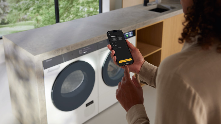Frau bedient eine Waschmaschine und einen Trockner mit einem Smartphone. Im Hintergrund ist ein Fenster und ein Regal zu sehen.