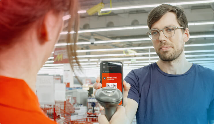 Ein Mann mit Brille hält ein Smartphone mit QR-Code und einen Scanner in einem Supermarkt. Eine Frau steht vor ihm.