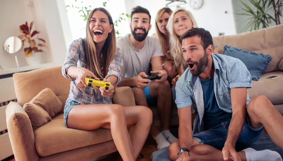 Fünf Personen sitzen auf einer Couch und spielen Videospiele. Eine Frau hält einen gelben Controller. Ein Mann sitzt auf dem Boden.