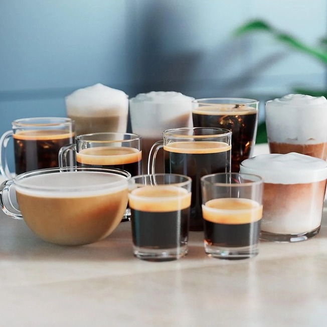 Elf verschiedene Kaffeegetränke in Gläsern und Tassen werden gezeigt, die sich in Größe, Schichtung und Milchschaum unterscheiden.