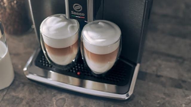Zwei Gläser mit Latte Macchiato stehen auf einer Kaffeemaschine. Die Getränke haben drei Schichten: Milch, Kaffee und Schaum.