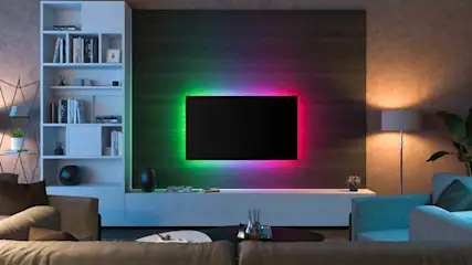 Ein Wohnzimmer mit Sofa, Tisch und Fernseher mit stimmungsvoller RGB Beleuchtung hinter dem TV und Lampen mit warmen Licht.