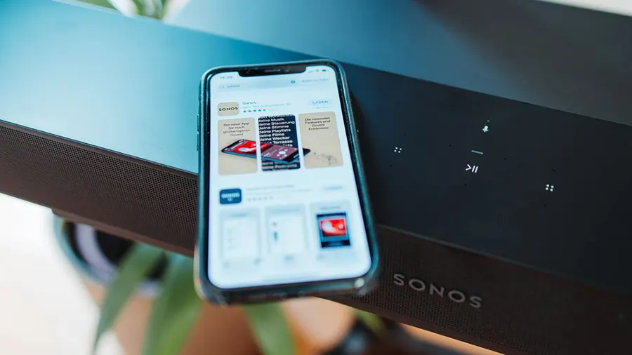 Ein Smartphone zeigt eine App, liegend auf einer schwarzen Sonos Soundbar mit Bedienfeld.