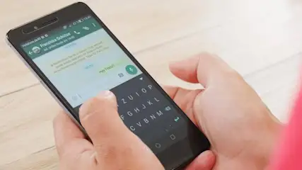 Hände halten ein Smartphone mit geöffnetem Chat und Tastatur auf einem Holztisch. Textnachrichten sind sichtbar.