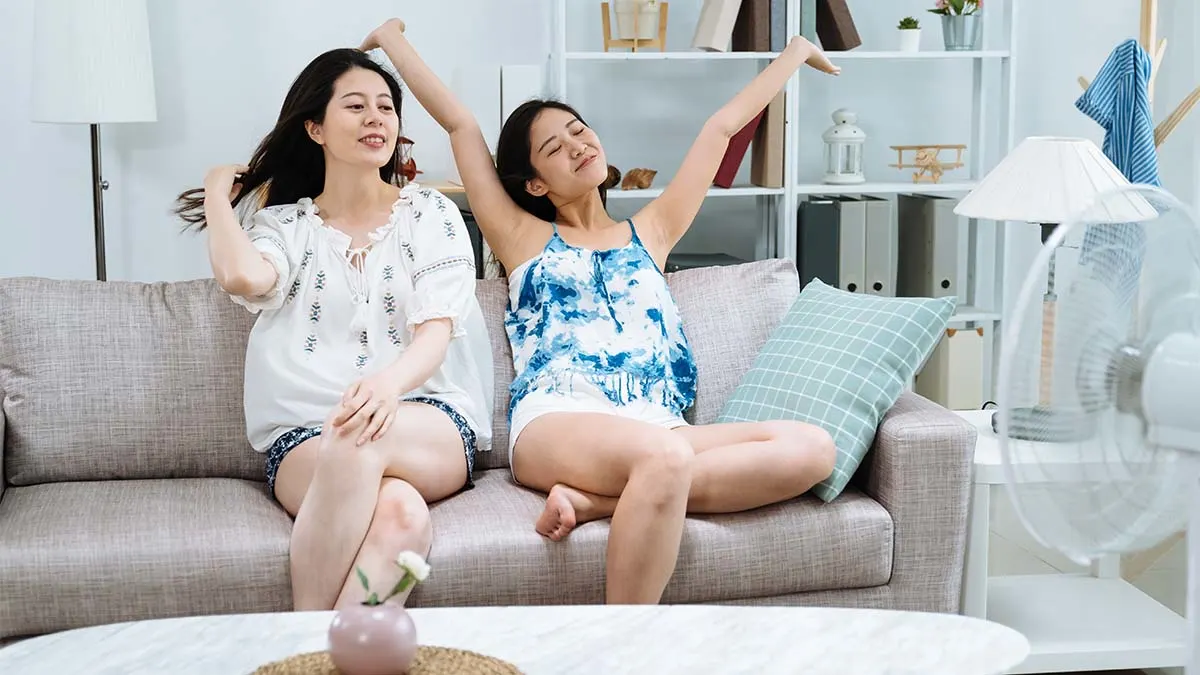 Zwei Frauen kühlen sich auf dem Sofa vor einem laufenden Standventilator ab.