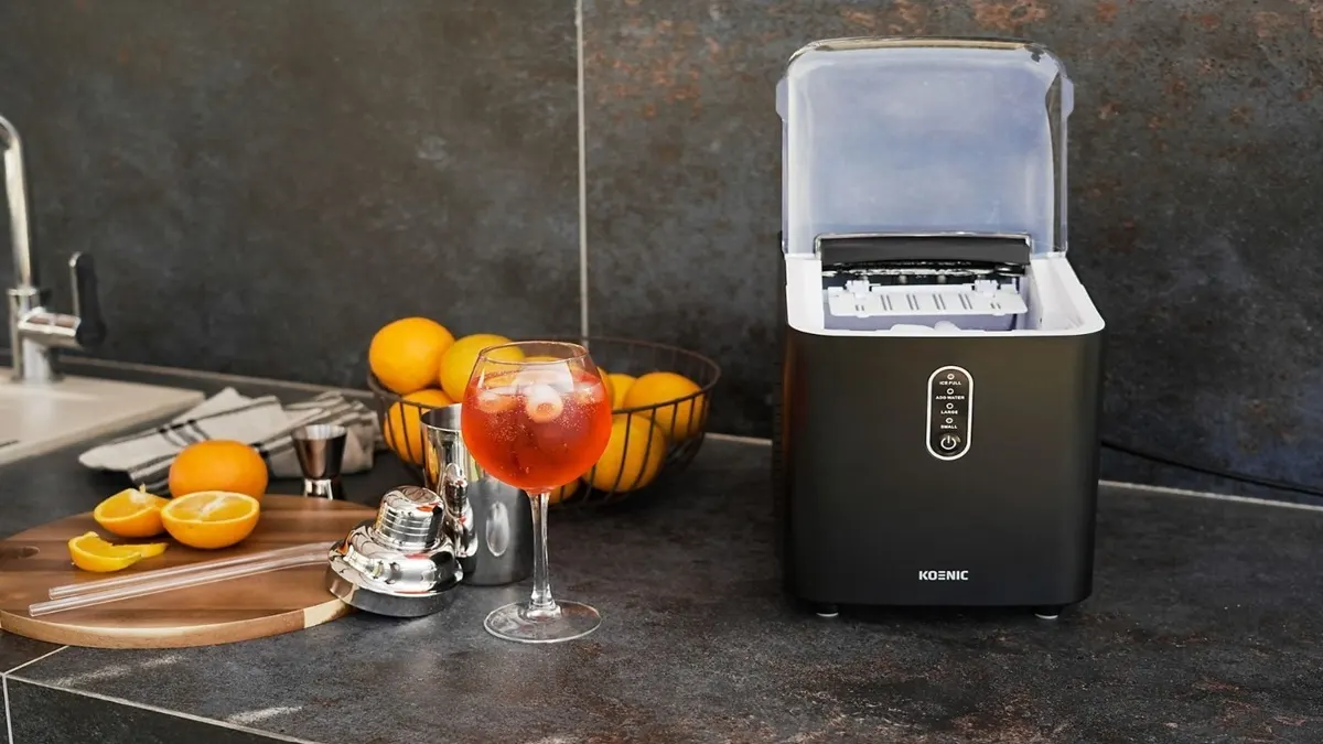 Der KOENIC KIC 8124 B Eiswürfelbereiter steht in einer Küche mit dunkler Arbeitsplatte. Daneben befinden sich ein Cocktailglas sowie Orangen in einem Korb und auf einem Schneidebrett.
