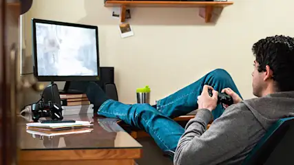 Mann spielt Videospiel mit Controller vor Monitor an Schreibtisch mit Büchern und Stiften in einem Zimmer.