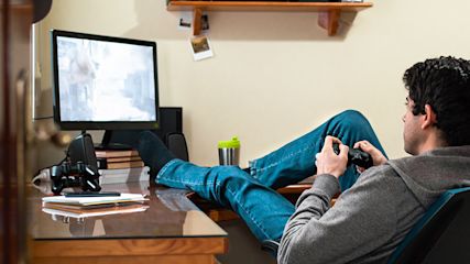 Junge Mann zockt am PC mit einem PlayStation-Controller. Er lehnt sich zurück und hat die Beide auf dem Schreibtisch.