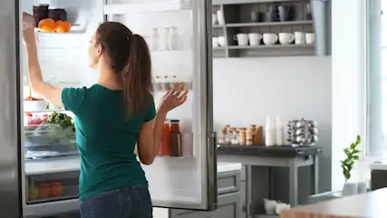 Eine Frau in einem grünen Hemd steht vor einem offenen Kühlschrank voller Lebensmittel. Die Küche ist hell und modern.