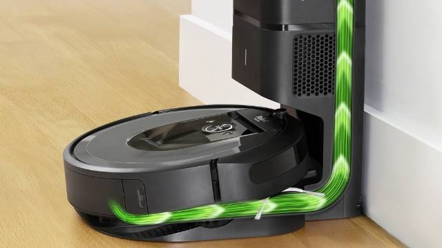 Ein schwarzer Staubsaugerroboter dockt an einer schwarzen Ladestation neben einer weißen Wand auf einem Holzboden an.