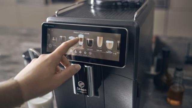 Eine Hand berührt den Bildschirm einer schwarzen Kaffeemaschine, auf dem Getränkeoptionen angezeigt werden.
