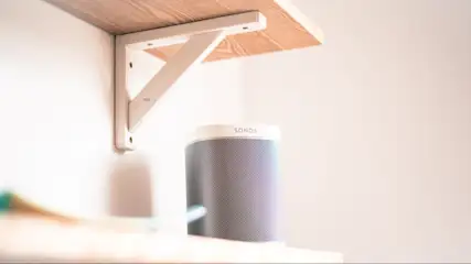 Ein Regal aus hellem Holz mit weißer Halterung an einer weißen Wand. Ein grauer Sonos-Lautsprecher steht auf dem Regal.