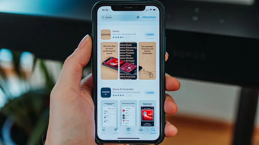 Eine Hand hält ein Smartphone, das den App Store mit der Sonos App zeigt. Der Hintergrund ist unscharf.