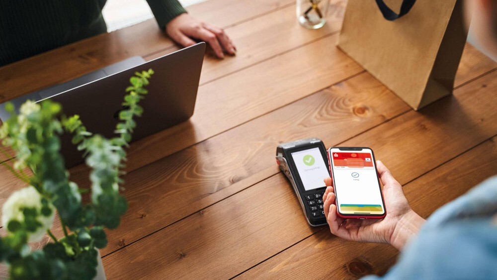 Holztisch mit Laptop, Pflanze, Papiertüte, Kartenzahlungsgerät und einer Person, die ein rotes Smartphone hält.