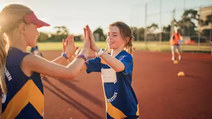 Zwei Mädchen in Sporttrikots klatschen sich auf einer roten Bahn ab. Im Hintergrund sind Bäume und eine Person zu sehen.