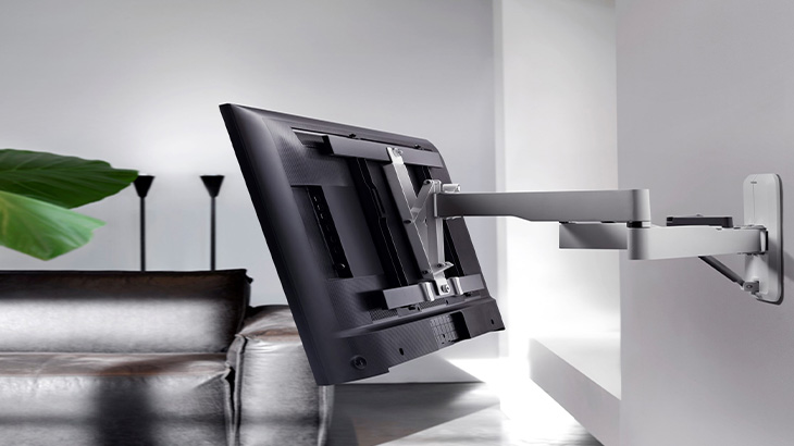 Ein schwarzer Fernseher ist an einer schwenkbaren, silbernen Wandhalterung befestigt. Im Hintergrund sind ein Sofa und Dekorationsgegenstände zu sehen.