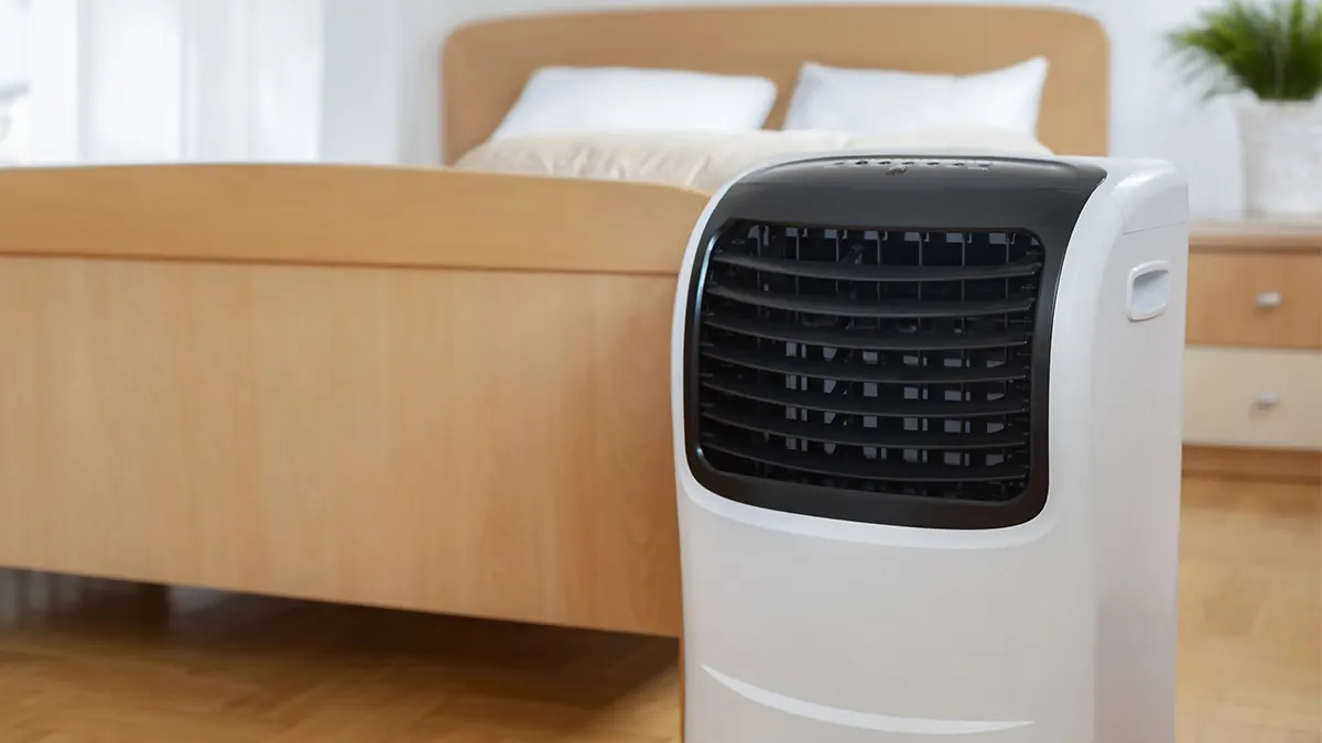Ein weißes mobiles Klimagerät steht im Schlafzimmer neben einem Holzbett.