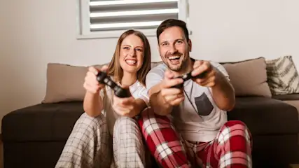 Ein Mann und eine Frau sitzen auf einem Sofa und halten Controller zum Videospielen. Sie tragen Pyjamas und lachen.