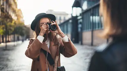 Eine Frau mit Hut fotografiert mit einer Kamera auf einer Straße, Gebäude und Bäume im Hintergrund.