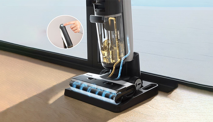Ein Staubsauger steht auf einem Holzboden vor einer Tür. Ein Finger zeigt auf den Griff. Der Staubsauger hat einen durchsichtigen Behälter.