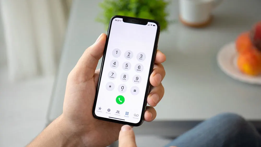 Eine Hand hält ein Smartphone mit geöffnetem Ziffernblock vor einem Tisch mit Obst und einer Tasse.