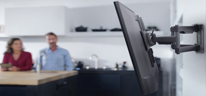 Ein an der Wand montierter Fernseher mit einer Halterung ist zu sehen. Im Hintergrund sitzen zwei Personen an einem Tisch.