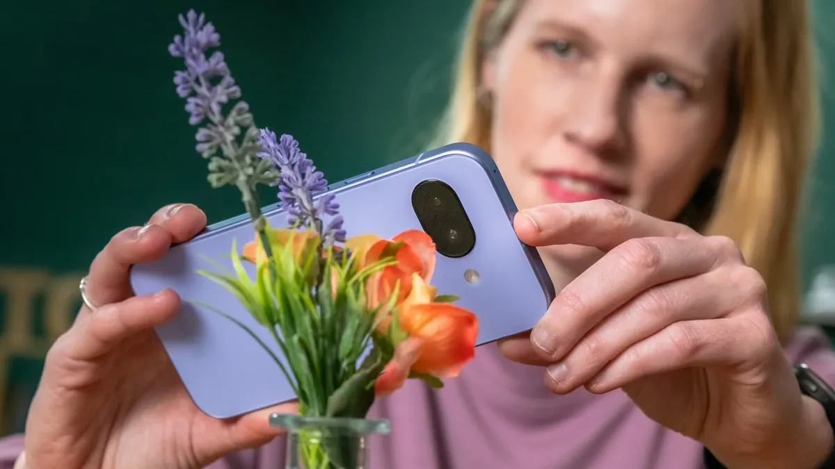Frau hält lila Handy mit Kamera über Blumen in Vase. Im Hintergrund ist eine grüne Wand zu sehen.