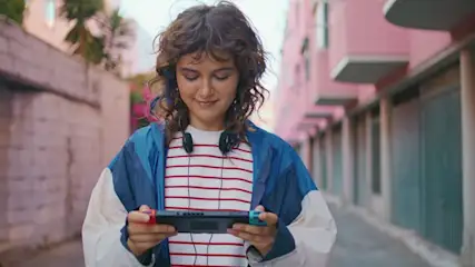 Eine Person hält eine Spielkonsole in einer Straße mit rosa Gebäuden. Sie trägt Kopfhörer und eine gestreifte Bluse.