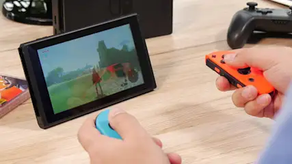 Ein schwarzer Nintendo mit Spiel auf dem Bildschirm und zwei Händen, die blaue und orangefarbene Controller halten.