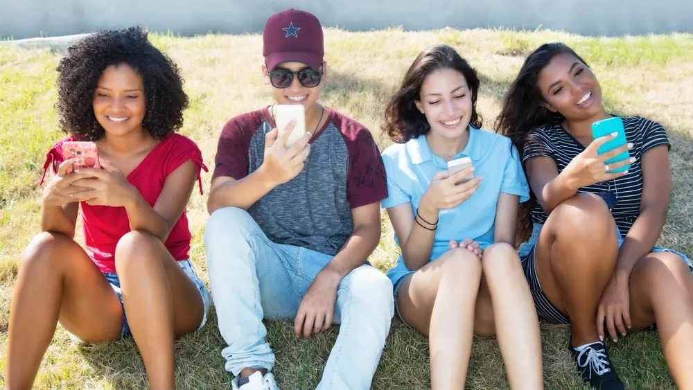 Vier junge Erwachsene sitzen auf Gras und schauen auf ihre Smartphones. Sie tragen lässige Kleidung und lächeln.