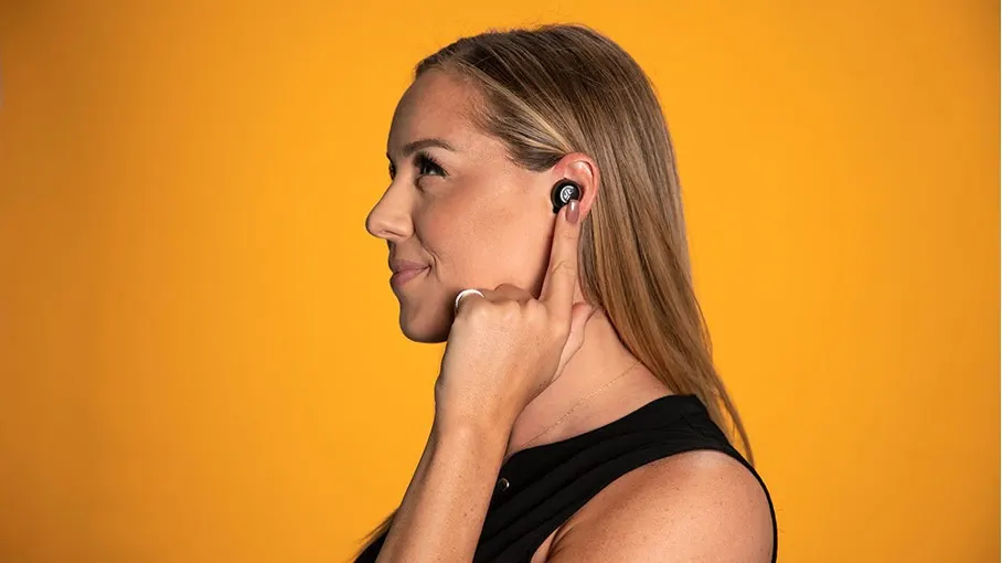 Profil einer Frau mit schwarzem Ohrhörer vor orangefarbenem Hintergrund. Sie berührt den Ohrhörer mit einem Finger.