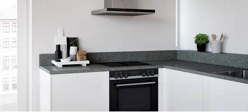 Helle Küche mit weißen Schränken, grauer Arbeitsplatte, schwarzem Herd und Dunstabzugshaube. Fenster im Hintergrund.