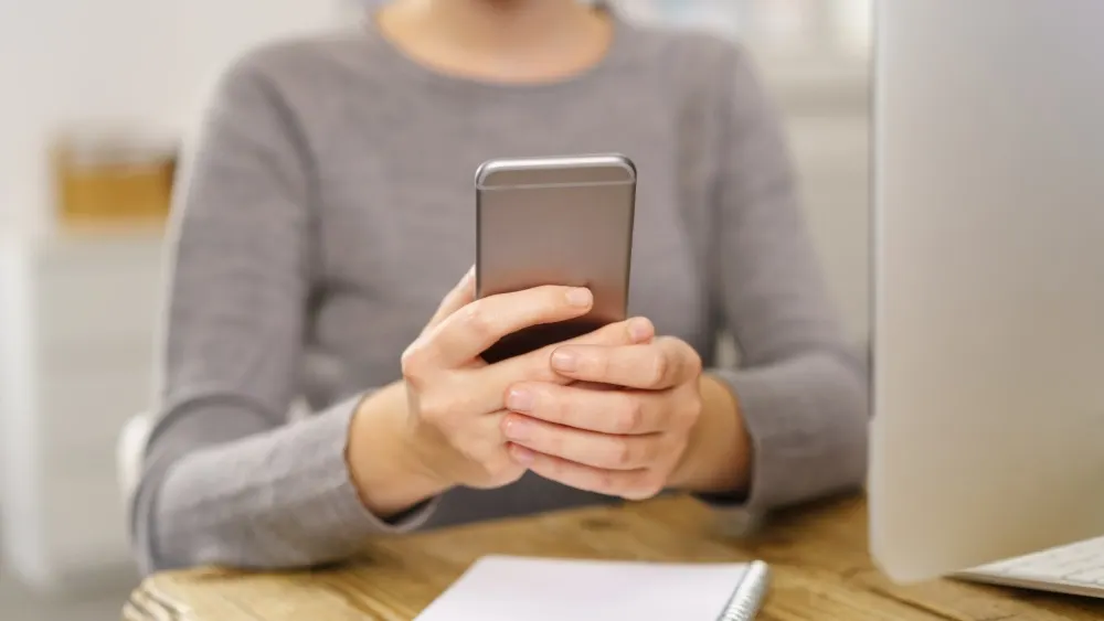 Eine Person im grauen Pullover hält ein graues Smartphone vor einem Computer und einem Notizblock auf einem Holztisch.