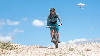 Ein Radfahrer mit Helm fährt einen Schotterweg hinunter, während eine Drohne am blauen Himmel fliegt.