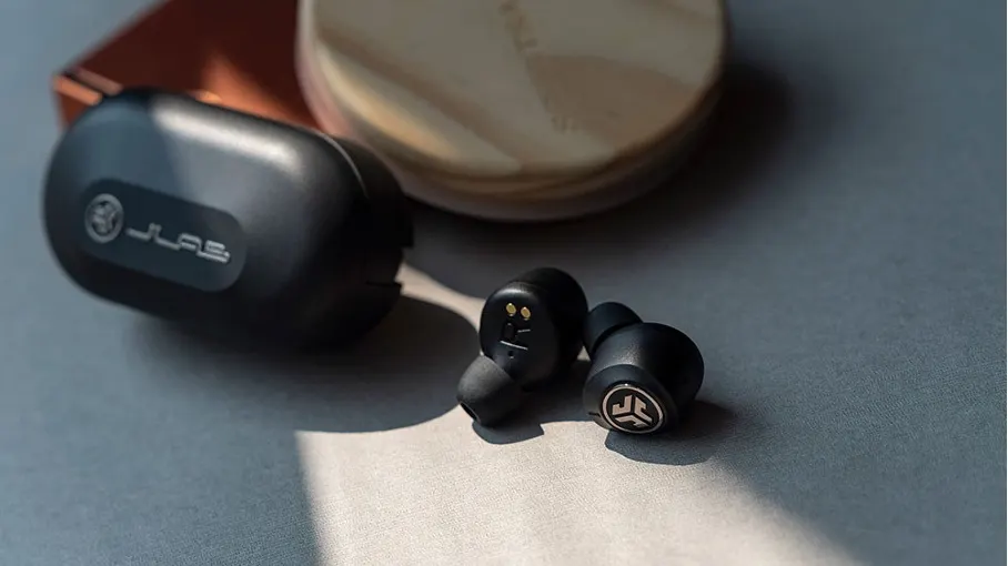 Schwarze JLab-Ohrhörer und ein Ladeetui liegen auf einer grauen Oberfläche, mit einem runden, hellen Gegenstand im Hintergrund.