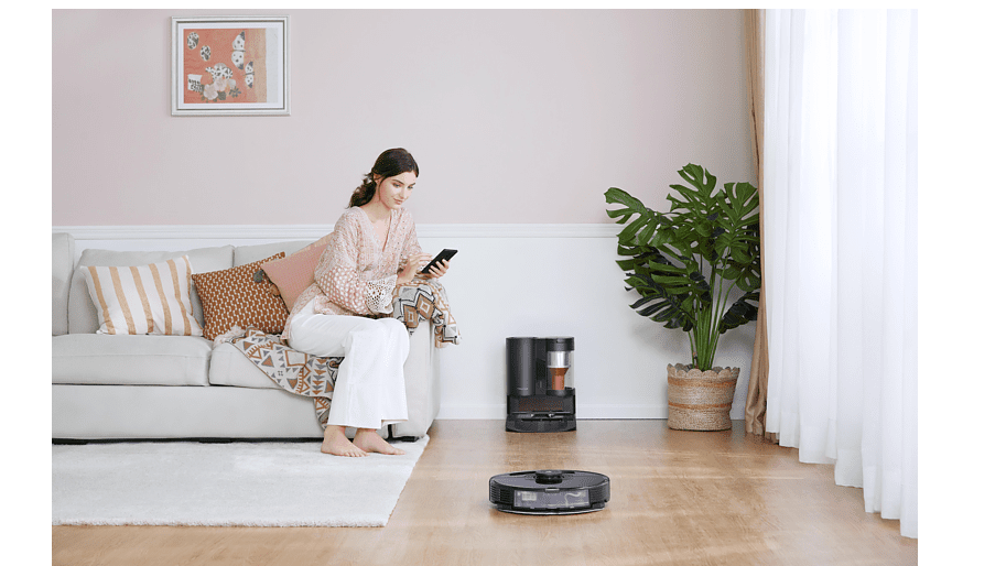 Eine Frau sitzt mit ihrem Smartphone auf der Couch. Vor ihr liegt ein Saugroboter