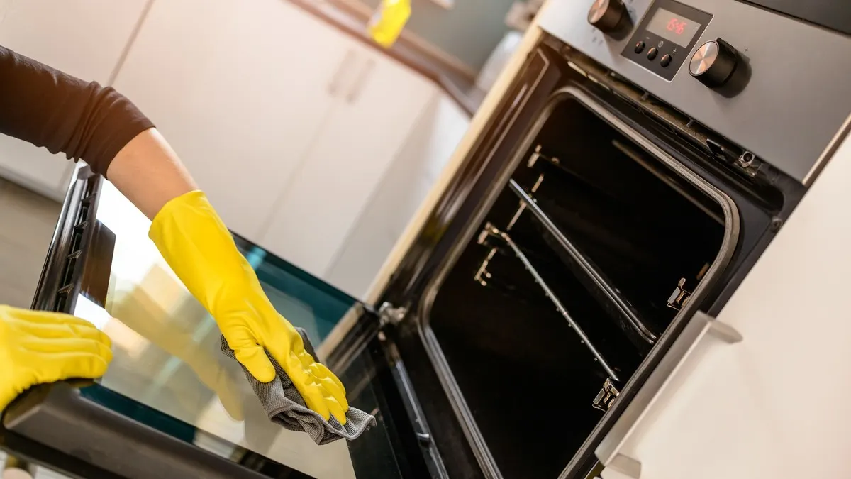 Eine Person mit gelben Handschuhen reinigt einen Ofen mit einem grauen Tuch in einer Küche.