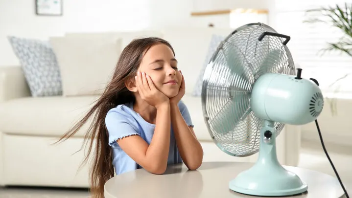 Ein Mädchen sitzt an einem Tisch vor einem laufenden, hellblauen Ventilator. Im Hintergrund sind ein Sofa und eine Pflanze.