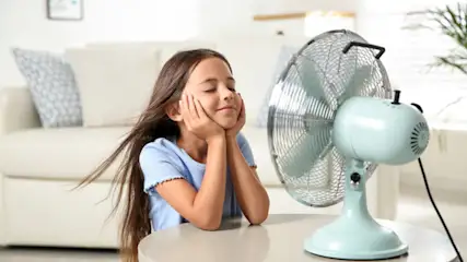 Ein Mädchen sitzt an einem Tisch vor einem laufenden, hellblauen Ventilator. Im Hintergrund sind ein Sofa und eine Pflanze.