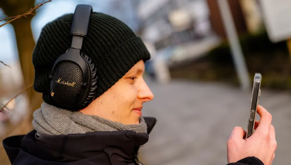 Ein Mann mit Mütze und Schal hört Musik mit schwarzen Marshall-Kopfhörern und hält ein Smartphone in der Hand.