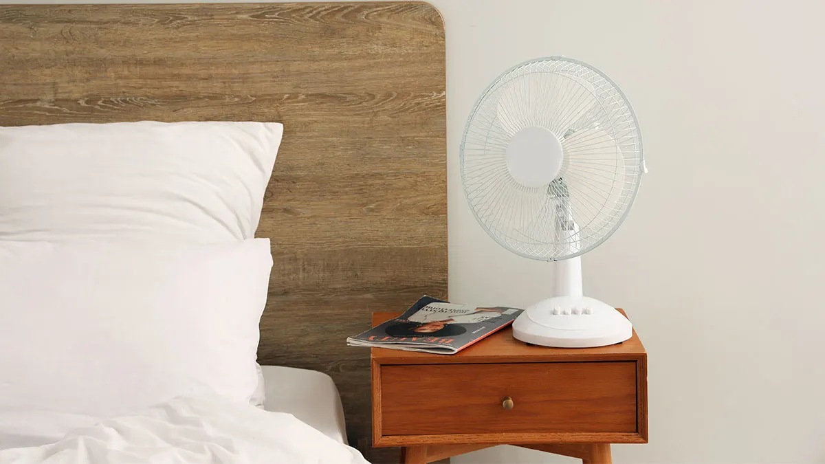 Weißer Tischventilator auf einem hölzernen Nachttisch neben einem Bett mit Kissen.
