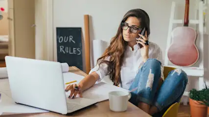 Eine Frau mit Brille telefoniert am Schreibtisch mit Laptop und Papieren. Im Hintergrund Tafel, Gitarre und Leiter.
