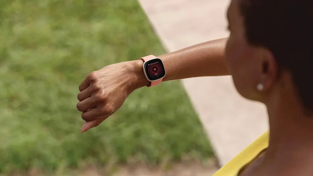 Eine Person, von hinten gesehen, trägt eine Smartwatch mit rosa Armband am linken Handgelenk. Gras ist im Hintergrund.