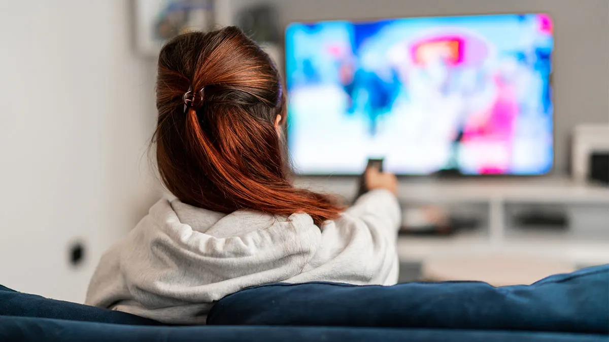 Eine Person mit langen Haaren sitzt auf dem Sofa und schaut fern. Sie hält eine Fernbedienung.