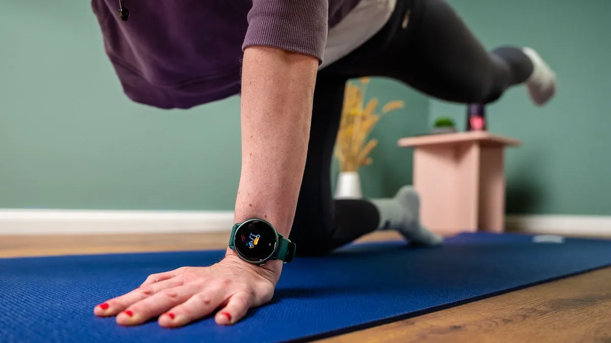 Eine Person trainiert auf einer blauen Matte mit einer Smartwatch am Handgelenk. Im Hintergrund steht ein Tisch mit Deko.