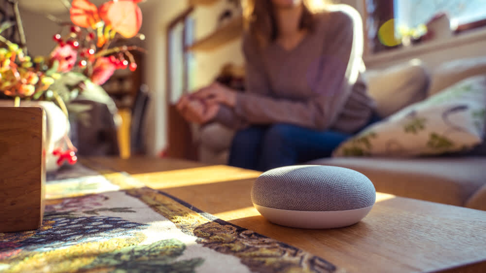 Ein grau-weißer Google Home Mini liegt auf einem Holztisch, im Hintergrund sitzt eine Frau auf dem Sofa.