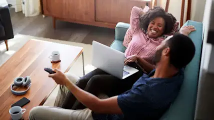 Ein Paar entspannt auf einem türkisfarbenen Sofa; sie mit Laptop, er mit Fernbedienung. Ein Tisch hält Kopfhörer und Tassen.